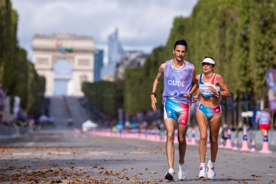Rosario Murcia-Gangloff, athlète paralympique française, court aux côtés de son guide lors du marathon des Jeux paralympiques de Paris 2024. En arrière-plan, l'Arc de Triomphe se dresse majestueusement, illustrant la grandeur de cet événement sportif.